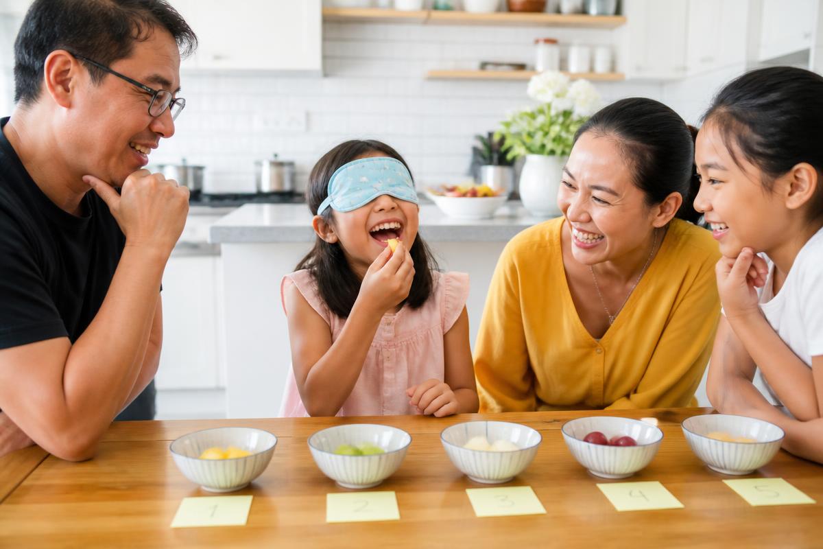 Niña con antifaz riendo mientras prueba una fruta en una cata a ciegas en casa